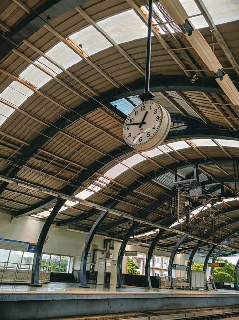 Photograph of Colliers Wood station platform interior showing a large round clock hanging from the ceiling with black framing and a glass canopy overhead. The platform has black metal benches, a ticket vending machine, and glass windows allowing natural light to illuminate the space. In the foreground, the edge of the platform is visible with protective barriers, and part of the railway tracks can be seen below. The station environment is clean and organized, with a curved roof design and lighting fixtures attached to the ceiling. The scene depicts a typical setting for a home relocation or furniture transport process, with ample space for loading and unloading in connection with house removals services by Colliers Wood Removals.