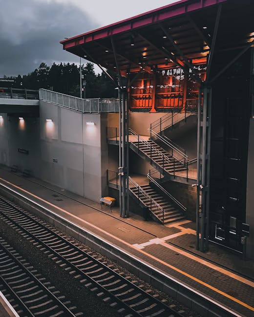 A photograph of an outdoor area near Colliers Wood station featuring a modern railway platform illuminated by artificial lighting. The platform has a textured yellow safety line along the edge, and the surroundings include railway tracks with black and silver rails, and gravel between the sleepers. Adjacent to the platform, a multi-level staircase with metal handrails and concrete steps provides pedestrian access between levels. The station building in the background has large windows, a red metal roof, and exterior walls made of glass and metal panels, reflecting an urban environment. The scene is captured during the evening or twilight, with dark clouds overhead and ambient lighting highlighting the station's structure. Colliers Wood Removals might utilise such a station environment for transport and home relocation logistics, ensuring efficient access and timing for furniture and boxes loading during removals.