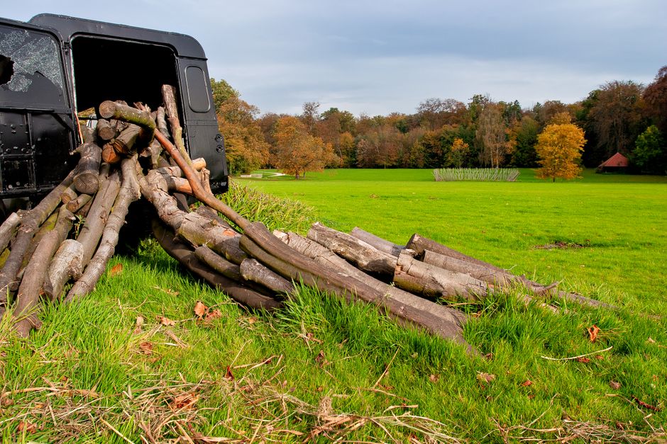 A black pickup truck parked on a grassy field with its rear liftgate open, displaying a pile of neatly stacked firewood consisting of logs of various sizes, some with bark still attached. Several logs are in the process of being loaded onto the truck, with a few partially lying on the ground next to it. The logs are arranged in an orderly manner, ready for transport or storage, and are wrapped in plastic covering. In the background, there is a wide expanse of green grass, a line of deciduous trees with autumnal foliage in shades of orange, yellow, and green, and a small red-roofed building partially visible among the trees. The scene is outdoors with soft, natural lighting, suggesting a clear or partly cloudy day, illustrating an outdoor staging area near a park or rural location, with the activity associated with home relocation or moving logistics by Colliers Wood Removals.