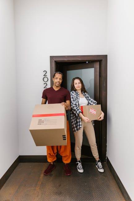 Two movers from Colliers Wood Removals are standing in a residential hallway near a doorway labeled with the number '202', each holding a packed cardboard box. The man on the left is dressed in a maro