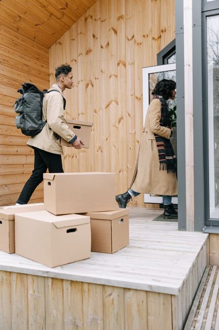Two movers from Colliers Wood Removals are standing in a residential hallway near a doorway labeled with the number '202', each holding a packed cardboard box. The man on the left is dressed in a maro